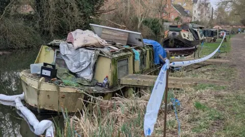 A badly run down narrowboat is covered in moss.  It is listing slightly alongside the towpath and a white plastic tap has been tied between metal stakes to cordon-off the boat from the towpath.