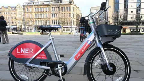 A grey and red e-bike parked in a paved city centre square on a sunny day. A man cycles away in the background