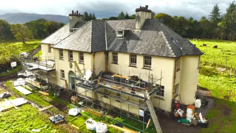 A drone shot from above of Ulva House, a run down two-storey mansion with scaffolding and building materials lying all around. Its idyllic setting is visible though, with grass and trees surrounding it and mountains in the distance