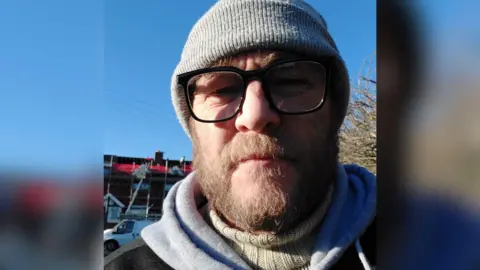 Scott Brooks, a man in his late 40s with a brown-haired beard and glasses, wearing a beanie hat.