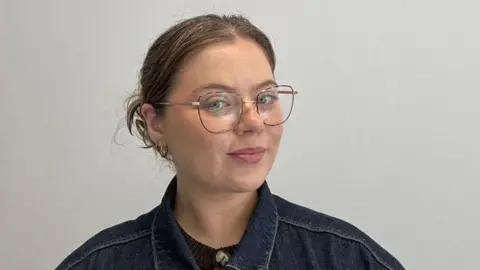 Maeve Schaffer Woman with brown hair, glasses and a denim jacket.
