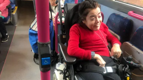 Joanne Sansome dark brown haired woman wearing a red jumper and sitting in a wheelchair in a wheelchair space on a bus 