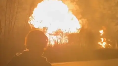 BBC A man stands in the foreground as a fireball erupts at the Ryazan oil refinery