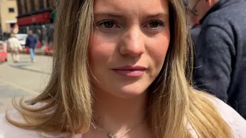 Woman with long blonde hair stands on a pavement, she wears a pink top