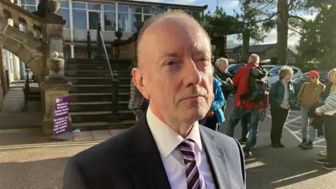 George Torr/BBC Head and shoulders shot of Dave Ratchford - he is wearing a black suit and a purple and pink tie.