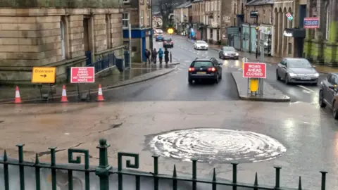 A mini roundabout on a town high street. There are yellow diversion and red road ahead closed signs.