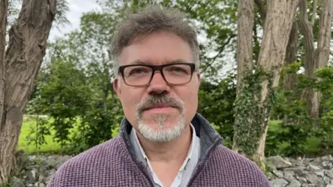A man with dark hair and a grey beard with dark rimmed glasses and a blue jumper with trees behind him