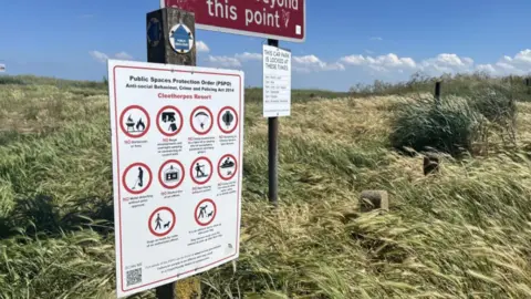 Lara King / BBC A white rectangular sign with red circles and black writing. Within the circles are warnings about what people cannot do on the beach. The sign is on a post next to two other signs which stand in long grass. The sky is blue and there are white clouds