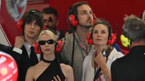 Reuters Actress Anya Taylor-Joy with boyfriend musician Malcolm McRae and actress Rebecca Ferguson with husband Rory St. Clair Gainer (centre) in the Ferrari garage during the race. All are wearing large red Ferrari headphones and looking at a screen.