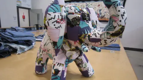 Alex Pope/BBC An elephant made from cloth, on a table with denim items on it. The elephant has a trunk, that is raised. 