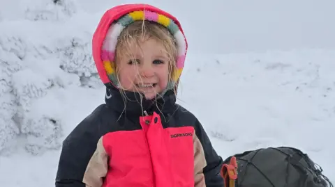 Five-year-old Faye is smiling at the camera in a snowy environment. She is wearing a bright pink jacket with black and beige sleeves. The hood is up over the top of the hood of a multi-coloured fleece. She has blonde hair which has bits of snow on it. She is standing next to a rucksack.
