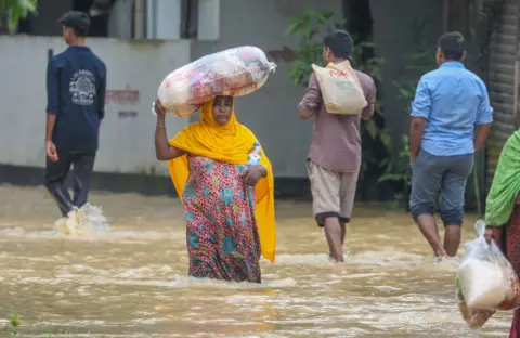     EPA-EFE/REX/Shutterstock Personas caminan entre las aguas inundadas en una zona afectada por las inundaciones en Burichong, distrito de Comilla, Bangladesh