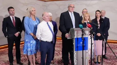 PA Media A group of politicians stand behind a podium at a press conference, with one man addressing microphones while others look on.