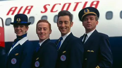 A female flight attendant stands proudly next to two male flight attendants and a male captain. They are all wearing navy blue blazers with an "AS" badge. A blue and white passenger jet is visible in the background with Air Scotia emblazoned on the side of it in red.