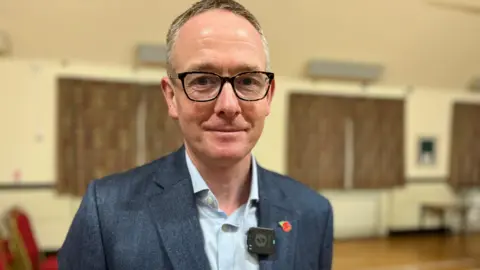 MP John Lamont looking at the camera. He is wearing glasses and a blue shirt with a blue suit jacket. He has a poppy pin in his lapel. He is in a hall and there are curtains drawn at windows behind him.