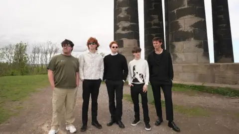 The band Swindled stand in front of Penshaw Monument in Sunderland and they look out at the horizon stoically. They are staring in different directions.