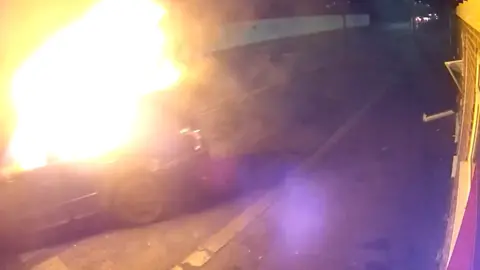A car on fire captured on CCTV - It is being moved from the front of a building.