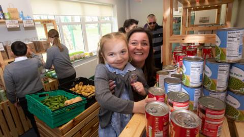 Woman holds child up in front of several tins of food