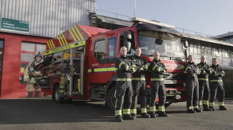 Six firefighters from the Kidlington Fire Station pose in front of their specialist vehicle outside their HQ. They have their arms folded in front of their chests. It is a sunny day.