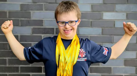 Joe Hughes, with two arms stretched out, with lots of yellow ribbons round his neck, he is smiling. He has on blue glasses and has short fair hair. He is standing by a brick wall and has on a blue T-shirt. 