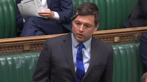 parliamentlive.tv A man with short brown hair, grey suit and blue shirt and tie stands in front of green leather seating. 