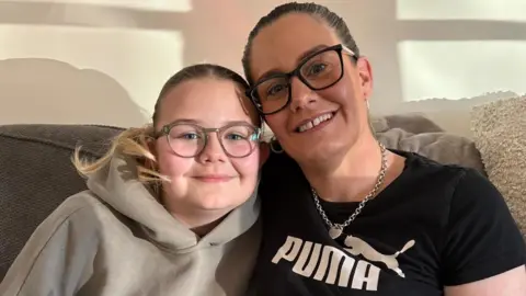 BBC A girl with her hair in a ponytail, wearing glasses and a beige hoodie, sits on a sofa next to a woman, also with her hair back and wearing glasses, wearing a silver necklace and earrings and a Puma T-shirt with a panther on it.