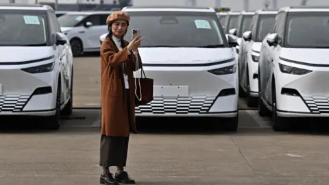 A woman takes a selfie in front X9 electric vehicles by Chinese EV manufacturer XPeng, are loaded on ship of the NYK line during a ceremony in the Port of Guangzhou, China's southern Guangdong province, on 22 February, 2025.