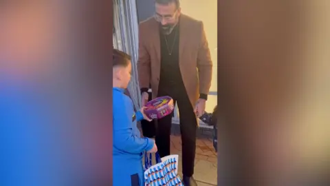 A boy in a blue tracksuit hands a box of chocolates to a man in the street