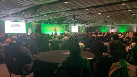 BBC A man standing on a stage with a green background, with a number of tables well attended by people watching the speech. 