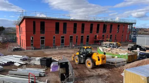 The building site of the University of Cumbria campus in Barrow. The building has no windows nor doors and is covered by red external panels. Building materials, a skip and a digger are in front of the building.