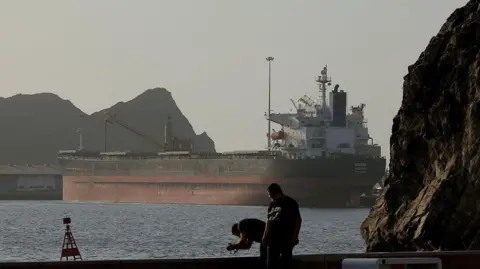 An oil tanker stands anchored between two rocky shores on a sunny evening. The image shows the Strait of Hormuz on 17 June