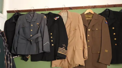 Jodie Halford/BBC A selection of vintage jackets and coats from the 1930s and 1940s hang on coat hangers on a green wall.