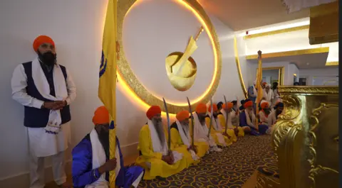 Guru Nanak Sikh Gurdwara About a dozen Sikh men sit on the floor against a wall in ceremonial dress and holding long, ceremonial swords with gold insignia on the wall behind them.
