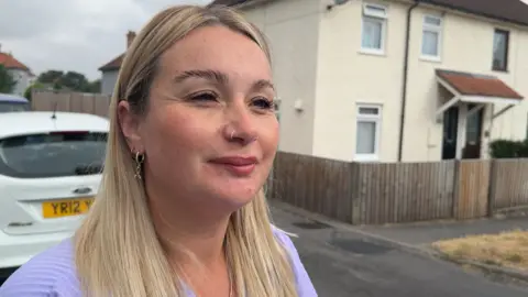 A woman with a nose piercing and long blonde hair hanging down over both shoulders squints at the camera through thick black eyelashes. She's wearing a purple blouse and stood in front of a white car.