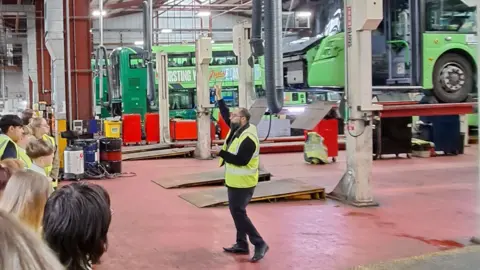 Ahead Partnership A man in a high vis jacket is stood in a bus depot and addressing a group of young students also in high vis. There's a green bus raised up on a platform behind him, with more buses in the background.