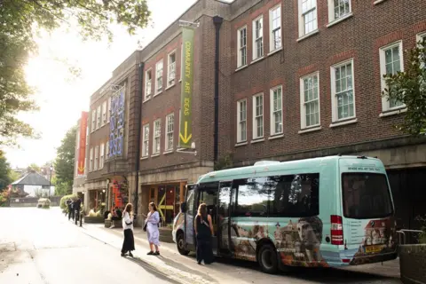 A blue shuttle bus outside a large building. There are passengers standing around the bus.