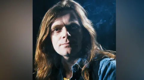 Guitarist Henry McCullough pictured in 1979. he has long light brown hair and is wearing a blue shirt