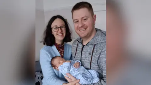 Contributed Brunette woman, wearing glasses and pale blue cardigan stands with sandy haired man wearing a grey hoodie. Both are smiling and he is holding a sleeping newborn baby in a pale blue babygrow.