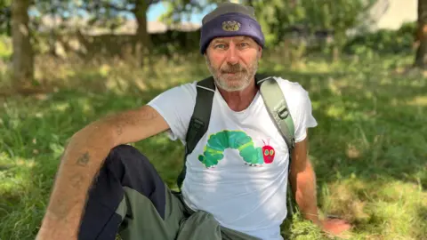 Steve England is sat on the grass, under a tree. He is wearing a navy beanie hat and a white t-shirt with a cartoon caterpillar on it. He is looking at the camera. He has a grey, short beard. 