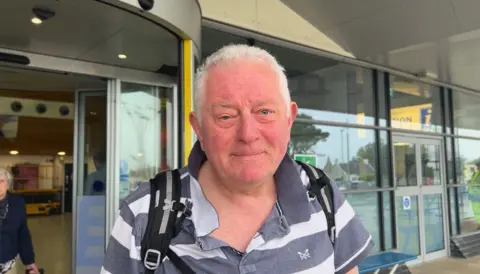 Phil Falla pictured in a grey and white striped polo shirt in front of airport terminal doors. He has short grey hair and is wearing a black backpack.