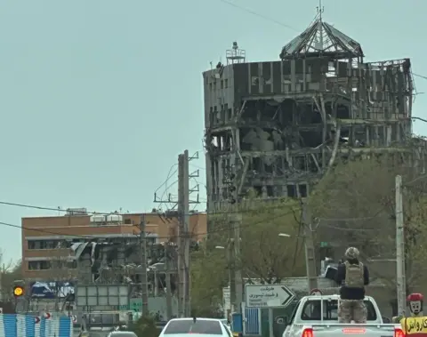 Supplied A figure in a military vest, helmet and fatigues standing in the back of a white pick-up truck, which is driving along a road in Tehran. A damaged building can be seen in the background.