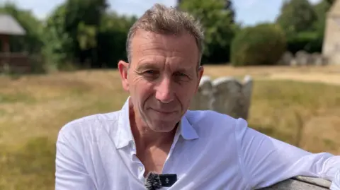 Michael Atherton has short brown hair and is wearing a white shirt whilst smiling.