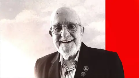 A bald elderly man with a beard smiles into the camera