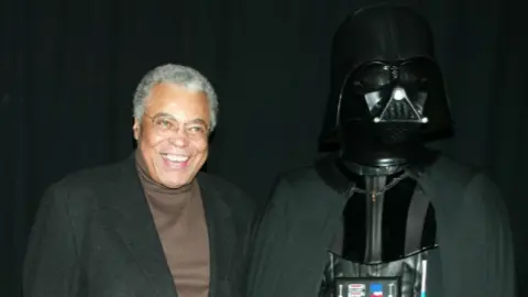 Getty Images James Earl Jones yang tersenyum berpose di sebelah kanan seseorang yang mengenakan kostum Darth Vader selama pemutaran perdana Star Wars: Episode II Attack of the Clones di New York pada tahun 2002. Jones mengenakan sweter cokelat dengan jaket hitam di atasnya.