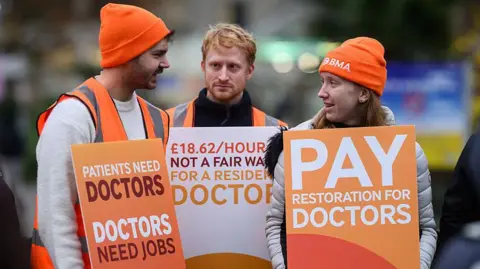 Getty Images Three resident doctors stand on a picket outside St Thomas' hospital in London at the start of a 5-day strike on 14 November 2025. They wear orange hats and hi-viz jackets and hold plackards reading "Patients need doctors, doctors need jobs" and "Pay restoration for doctors". 