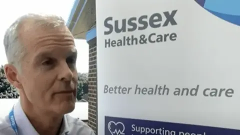 Stephen Lightfoot, a white man with silver hair, stands in front of a sign that reads "Sussex Health and Care - better health and care" 