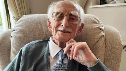 Man wearing glasses looks straight into the camera lens. He's smiling and wearing a white shirt with a blue and white spotted tie and grey cardigan
