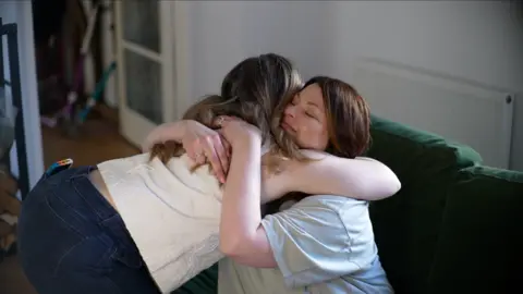Jamie Niblock/ BBC Liv and Lucy embrace in a hug. They are comforting each other after becoming emotional during an interview. Lucy is smiling, as she closes her eyes. 