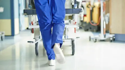 A person is pushing a hospital bed down a hospital corridor. The person is wearing blue trousers and white shoes. There are items on wheels on both sides of the corridor. The floor is an off white colour with a blue border. 