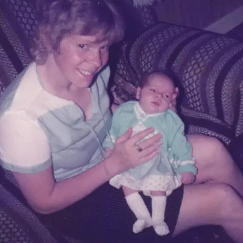 Family of Kirsty Wilkinson Cathy sits in an armchair with baby Kirsty. Cathy's hair is short and she wears a short sleeved green and white top and a dark skirt. Kirsty is dressed in a white spotted dress with a green cardigan over it and white knee length socks.